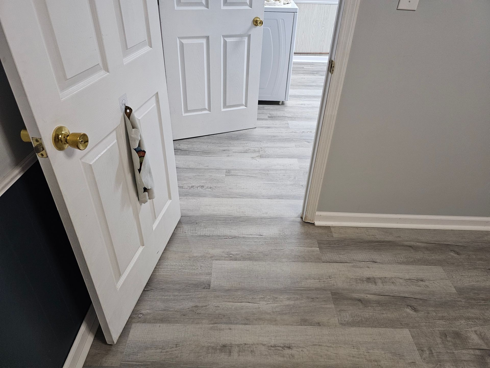A doorway opens to a room with grey wood-look flooring and multiple white doors.