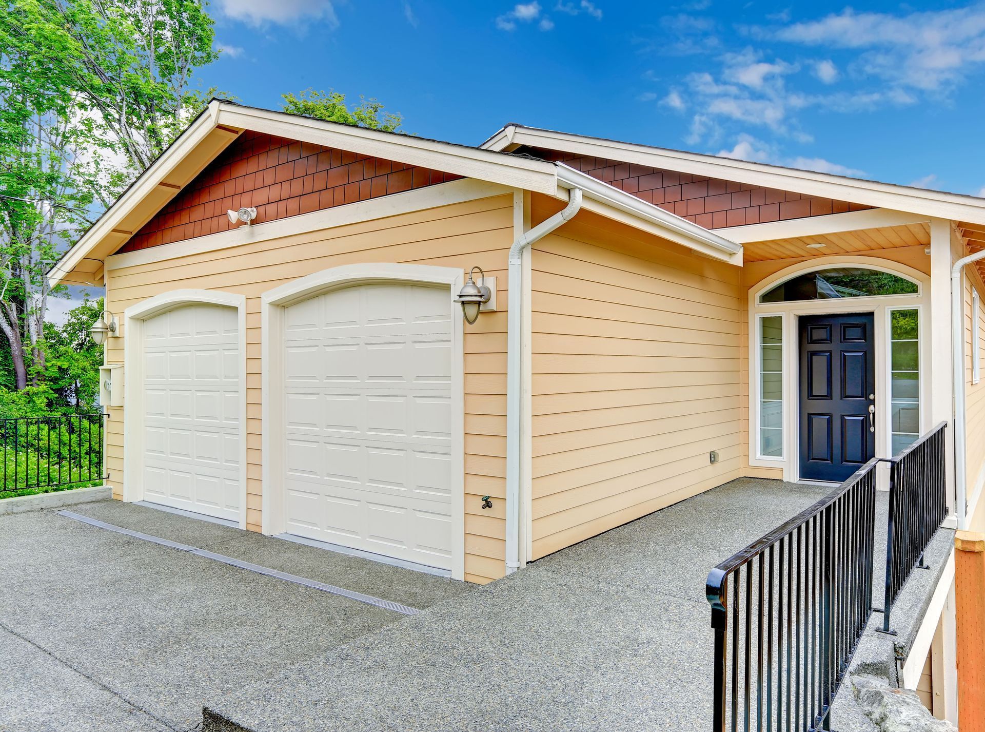 Two-car garage with white doors, tan siding, and a black railing beside a gravel driveway