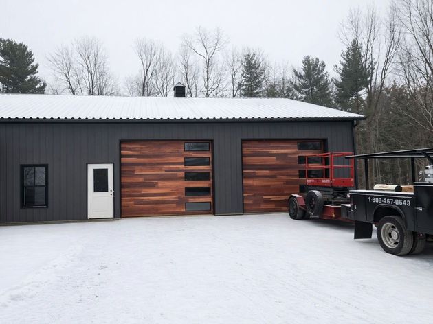Snowy yard with a dark garage and wooden doors, plus a red truck and trailer parked beside it.