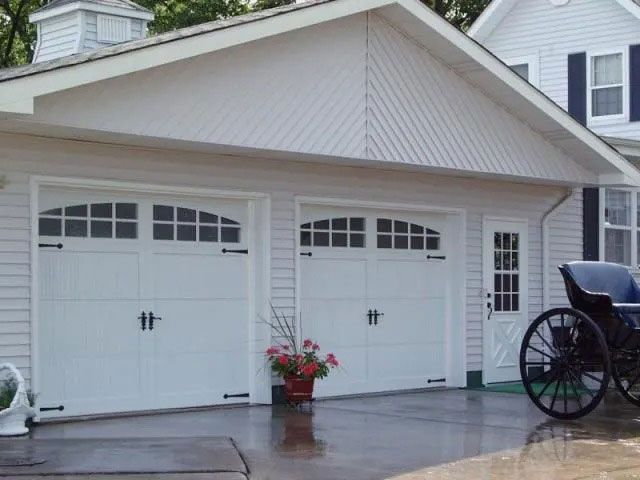 White garage doors with a flower pot beside them and a horse-drawn carriage wheel at right