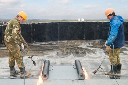 Two workers in hard hats inspect a rooftop membrane with tools and a torch.