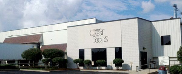 Exterior of a white Crest Foods warehouse building with trimmed shrubs and a parking lot.