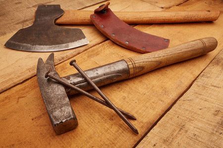 Three woodworking tools on a wooden table: an axe, a hatchet, and nails with a hammer.