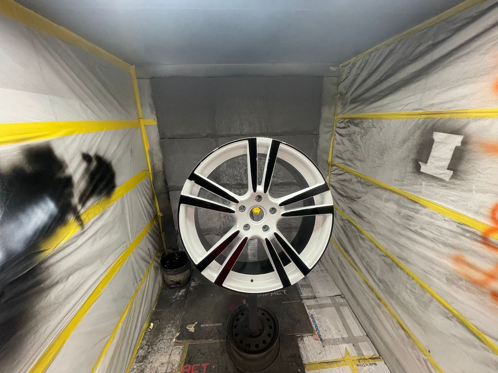 A white car wheel with black stripes being painted inside a spray booth with yellow tape.