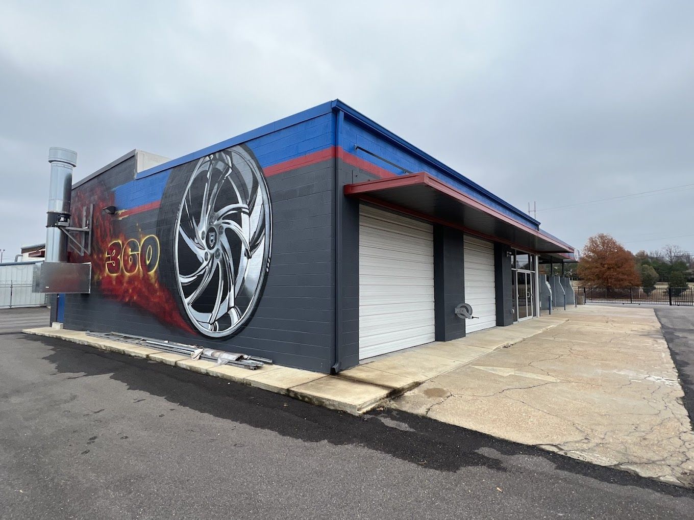 A building with a mural of a car wheel and tire. Gray, red, and blue exterior. Two garage doors.