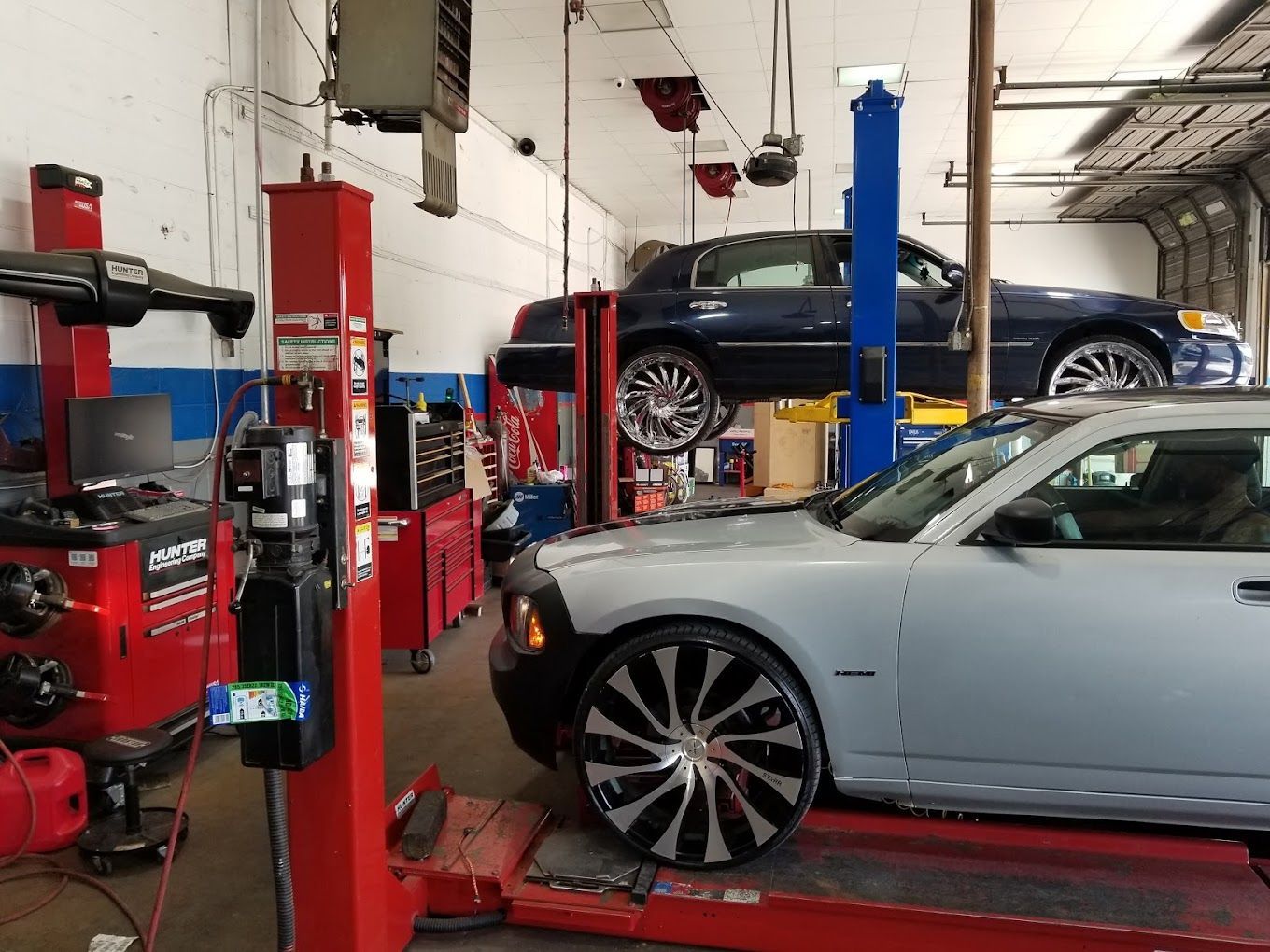 Car repair shop: two cars on lifts, gray Dodge Charger and blue sedan. Tools and equipment are visible.