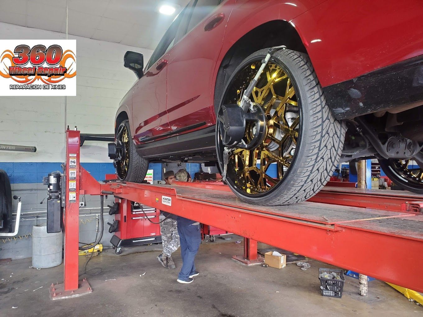 Red car on a lift in a repair shop, wheels are gold. A person is underneath the car.