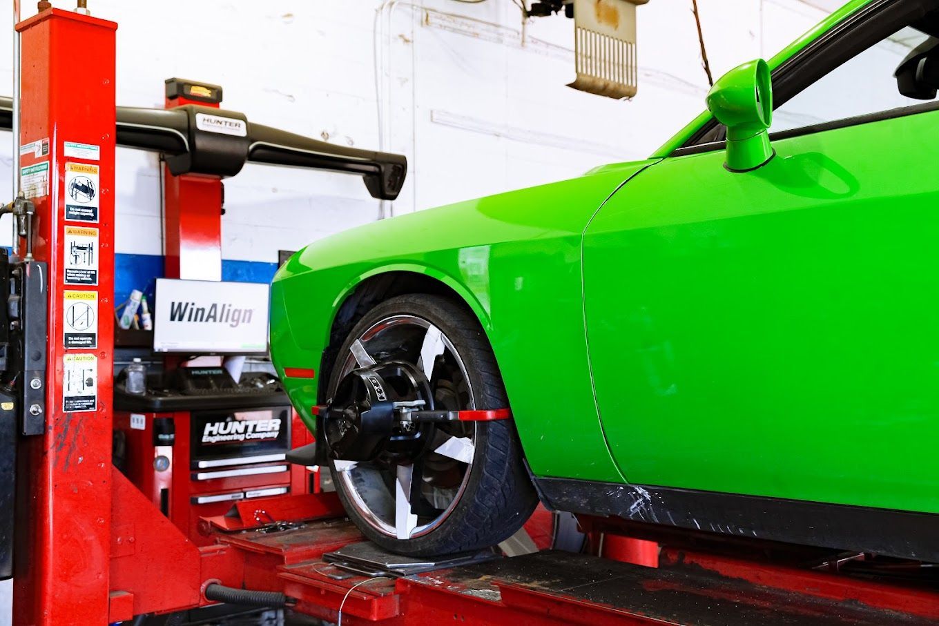 Green car on a lift being aligned in a garage. Red lift and tools visible.