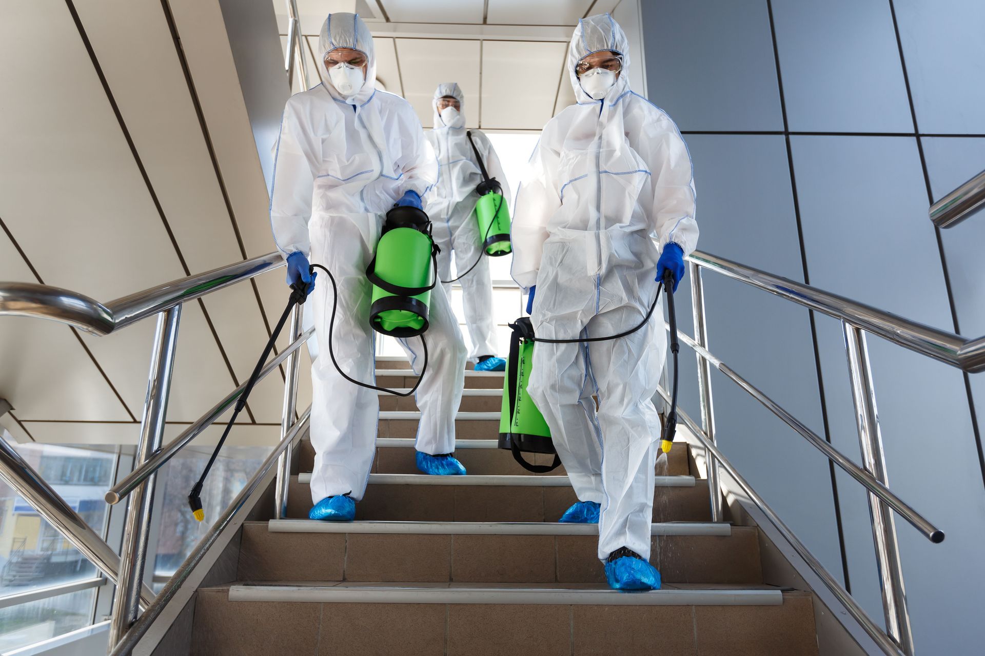 Three workers in white hazmat suits carrying green cleaning sprayers down a stairway