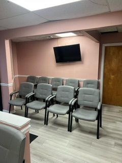 A row of chairs in a waiting room with a flat screen tv on the wall.