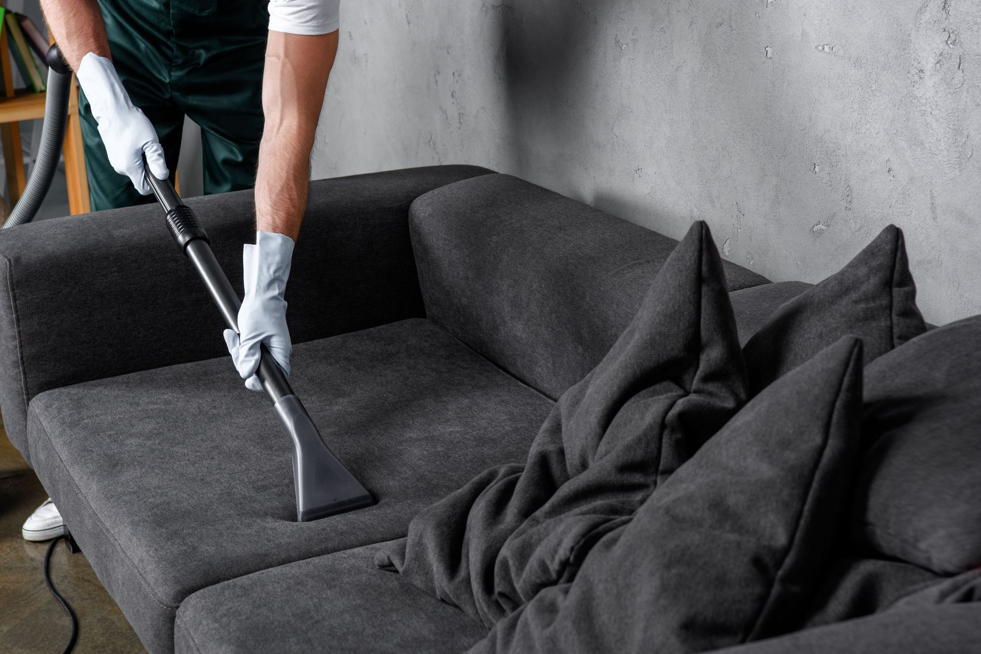 Person vacuuming a dark gray couch with a hose and nozzle, wearing gloves.