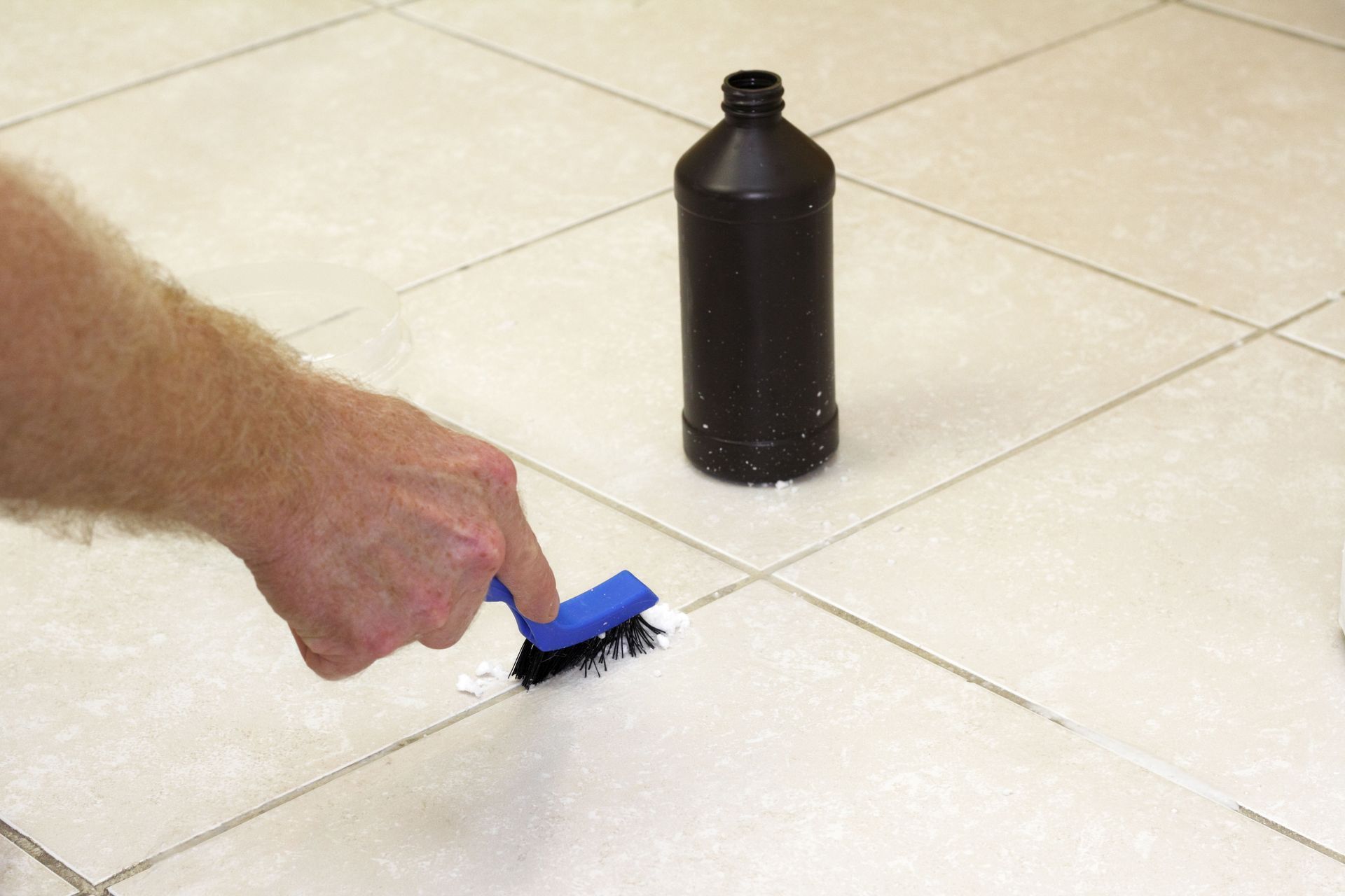 Person scrubbing grout with brush on tiled floor, black bottle nearby.