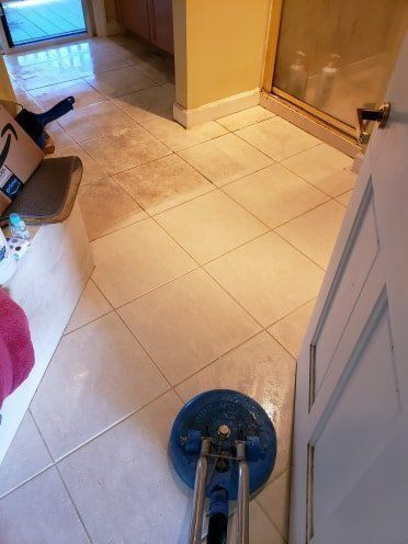 Floor being cleaned with a blue scrubbing machine in a bathroom with beige tiles and white door.
