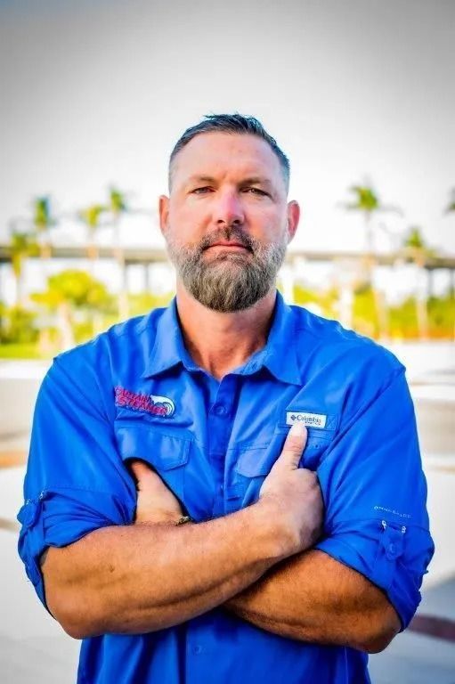 Man with beard in blue shirt, arms crossed, standing outdoors.