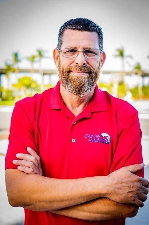Man with glasses and beard wearing a red polo shirt, arms crossed, standing outdoors.