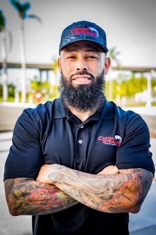Man with full beard, tattoos, wearing a logo cap and polo, arms crossed, smiling.
