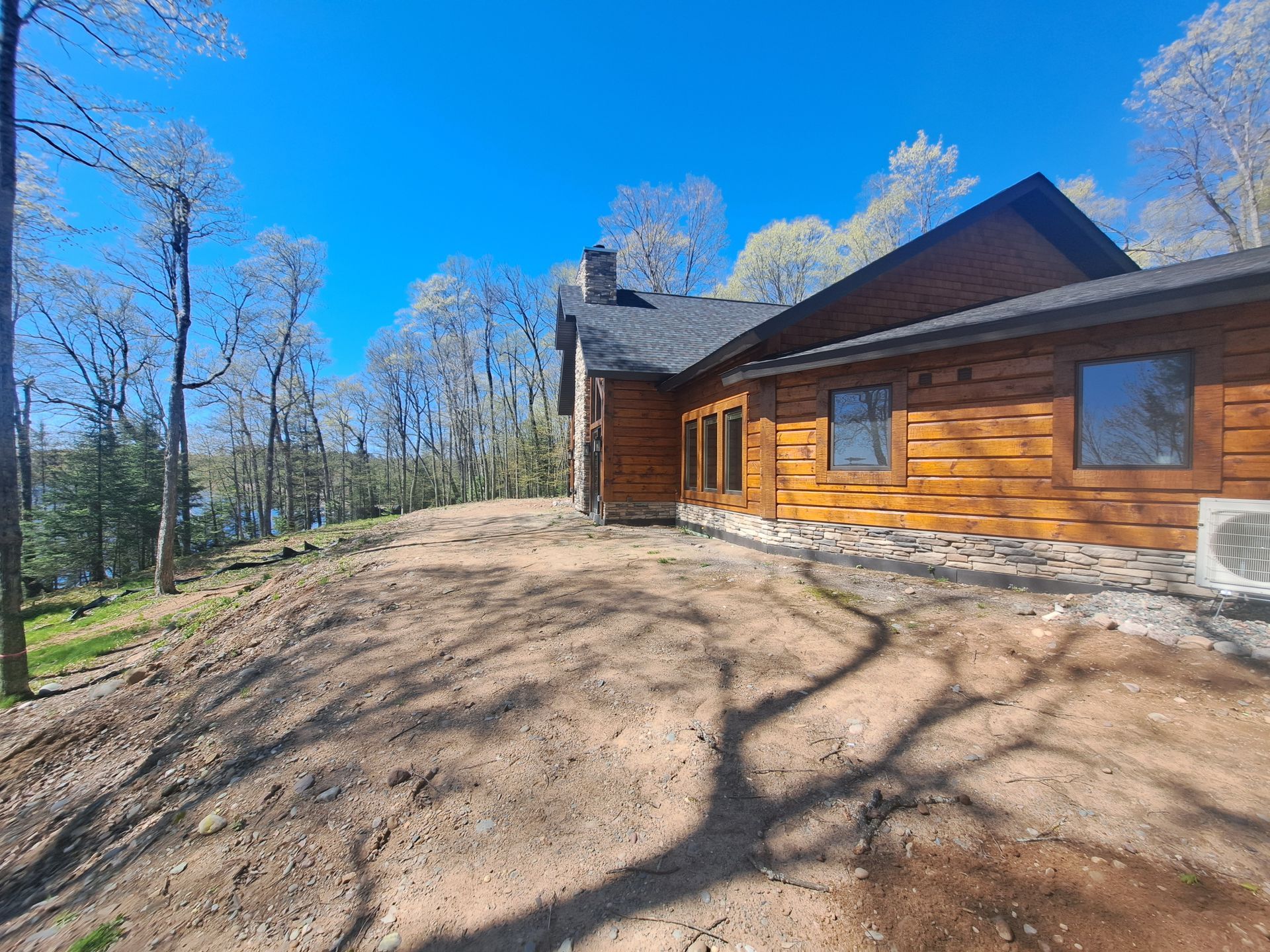 Log cabin with brown wood siding, on a hill with bare soil, trees, and blue sky.