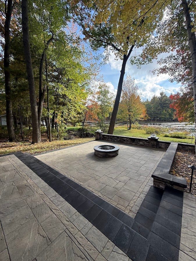 Outdoor stone patio with a fire pit, surrounded by trees and autumn foliage under a clear sky