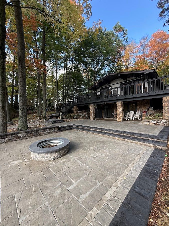 Stone patio with circular fire pit in front of a dark cabin among tall autumn trees.