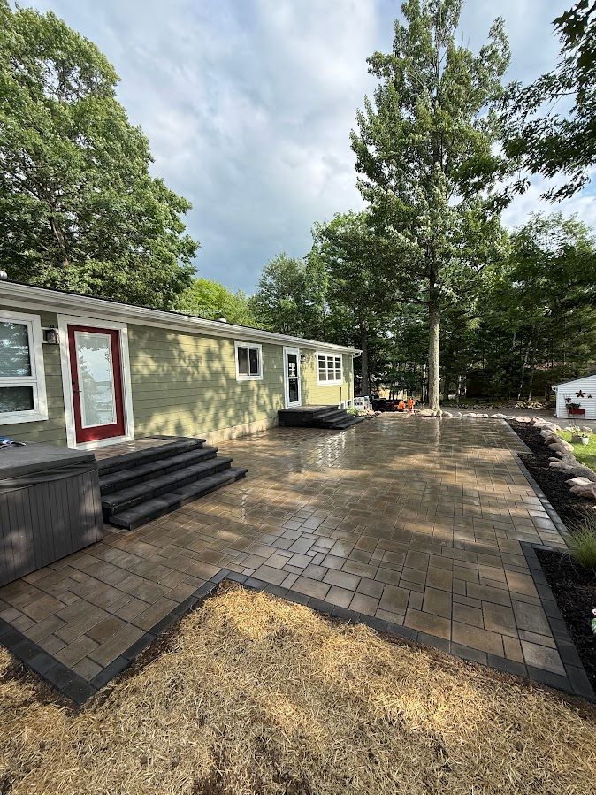 Backyard with new gray paver patio beside a house, black steps, and trees under a cloudy sky