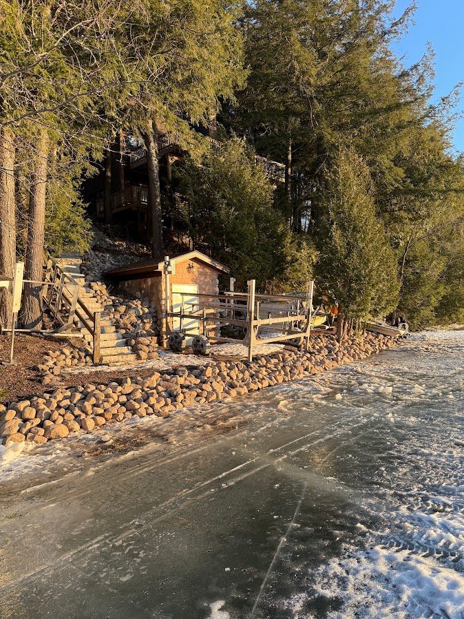 Rocky shoreline with a rustic cabin, wooden fence, and trees beside icy water.