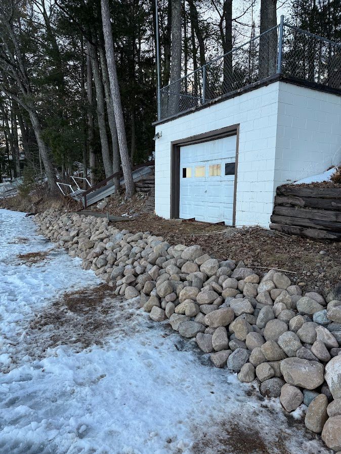 Snowy outdoor utility shed with a white garage door, stone retaining wall, and wooded hillside.
