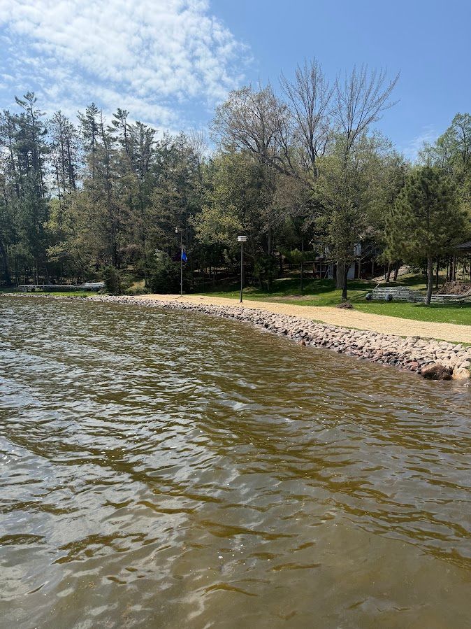 Rocky shoreline beside rippling water with trees and a small grassy park in the background