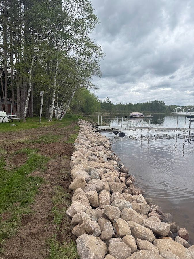Rock-lined shoreline beside a grassy path and trees, with calm water and a cloudy sky