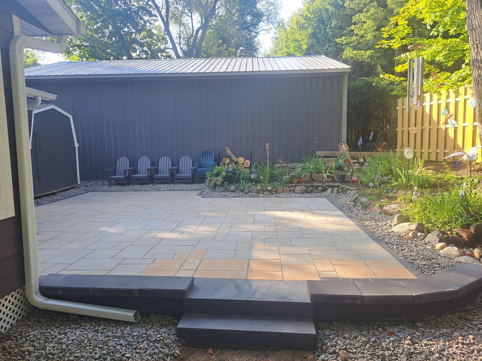 A paved patio with steps, gravel, and a shed in the background. Blue chairs are visible.