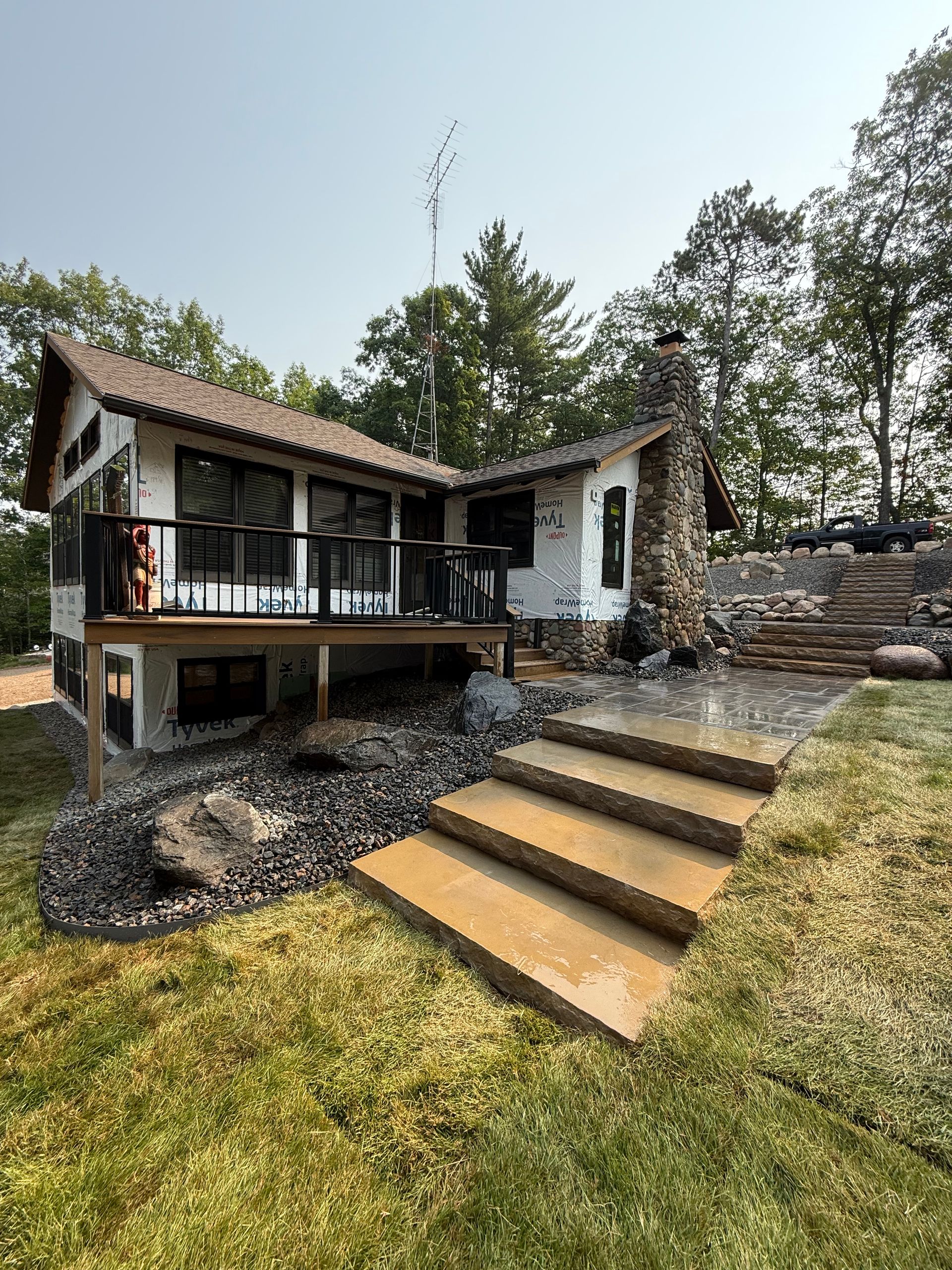 A white cabin with a deck, stone steps, and a chimney surrounded by green grass and trees.