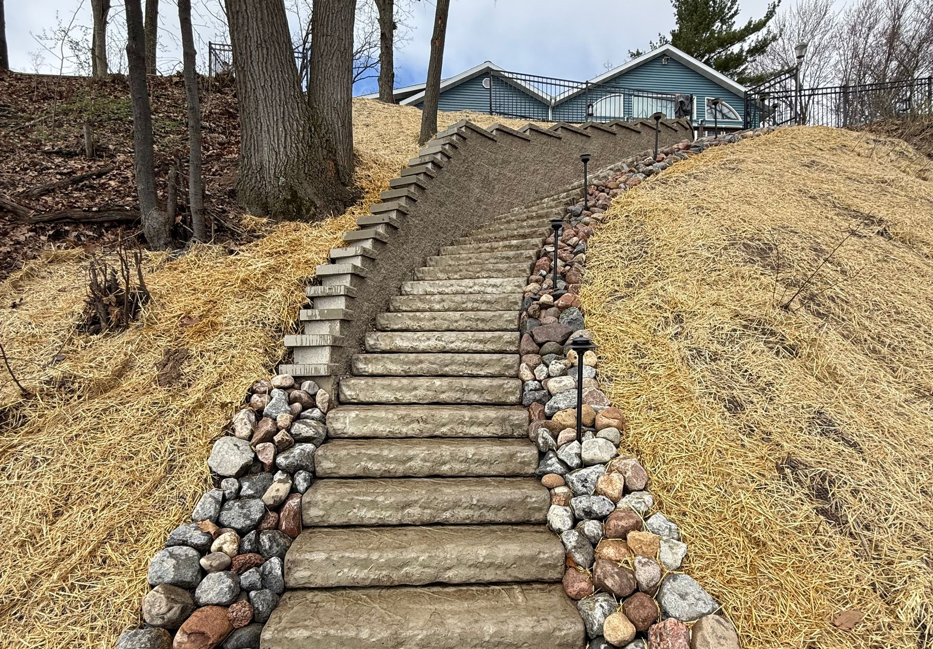 Stone steps lead uphill to blue houses on a hill, flanked by rock and mulch.