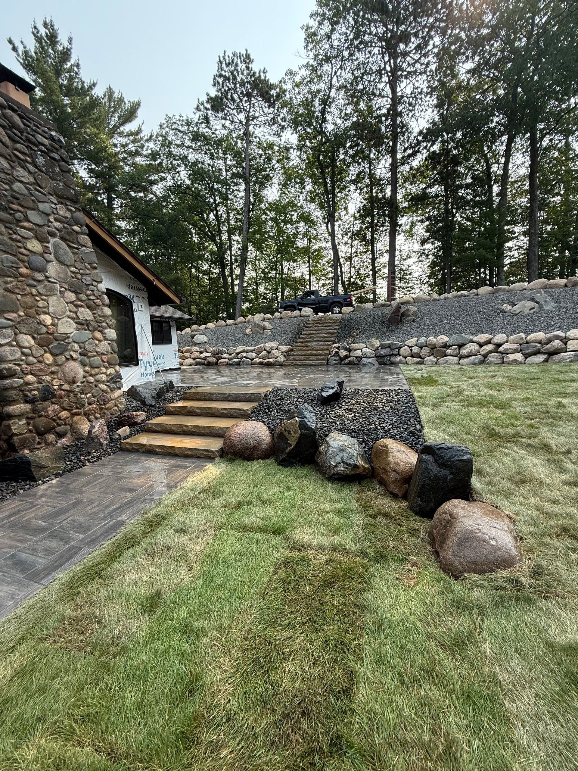 Stone house exterior with steps leading to a raised area lined with stones, rocks, and trees.