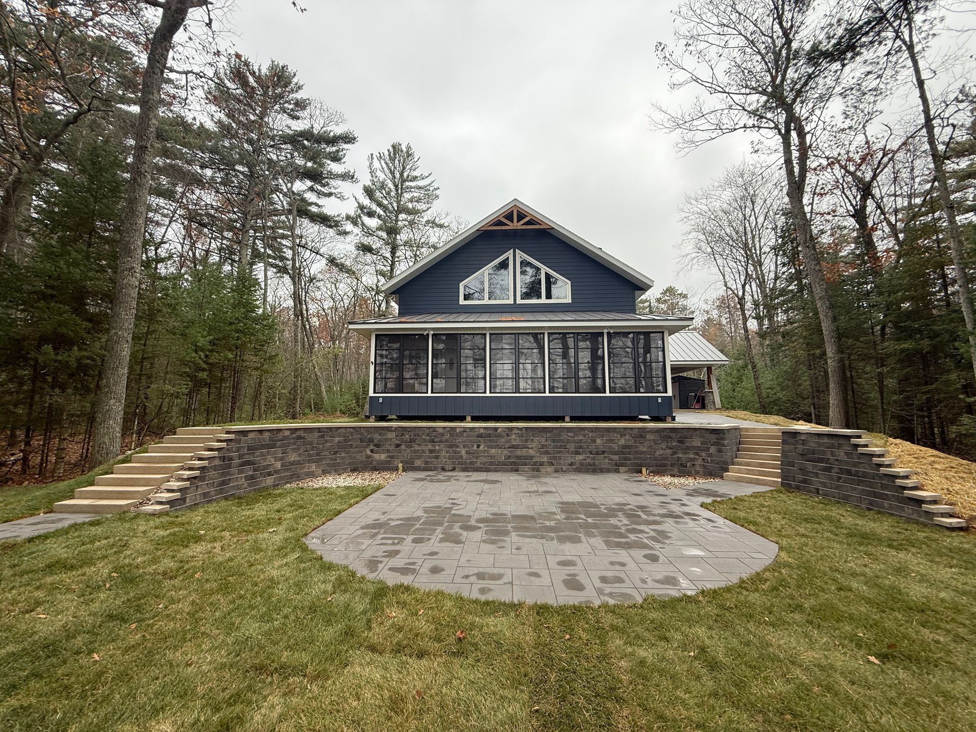 Blue house with screened porch, patio, and stone retaining wall set in a wooded area on a cloudy day.
