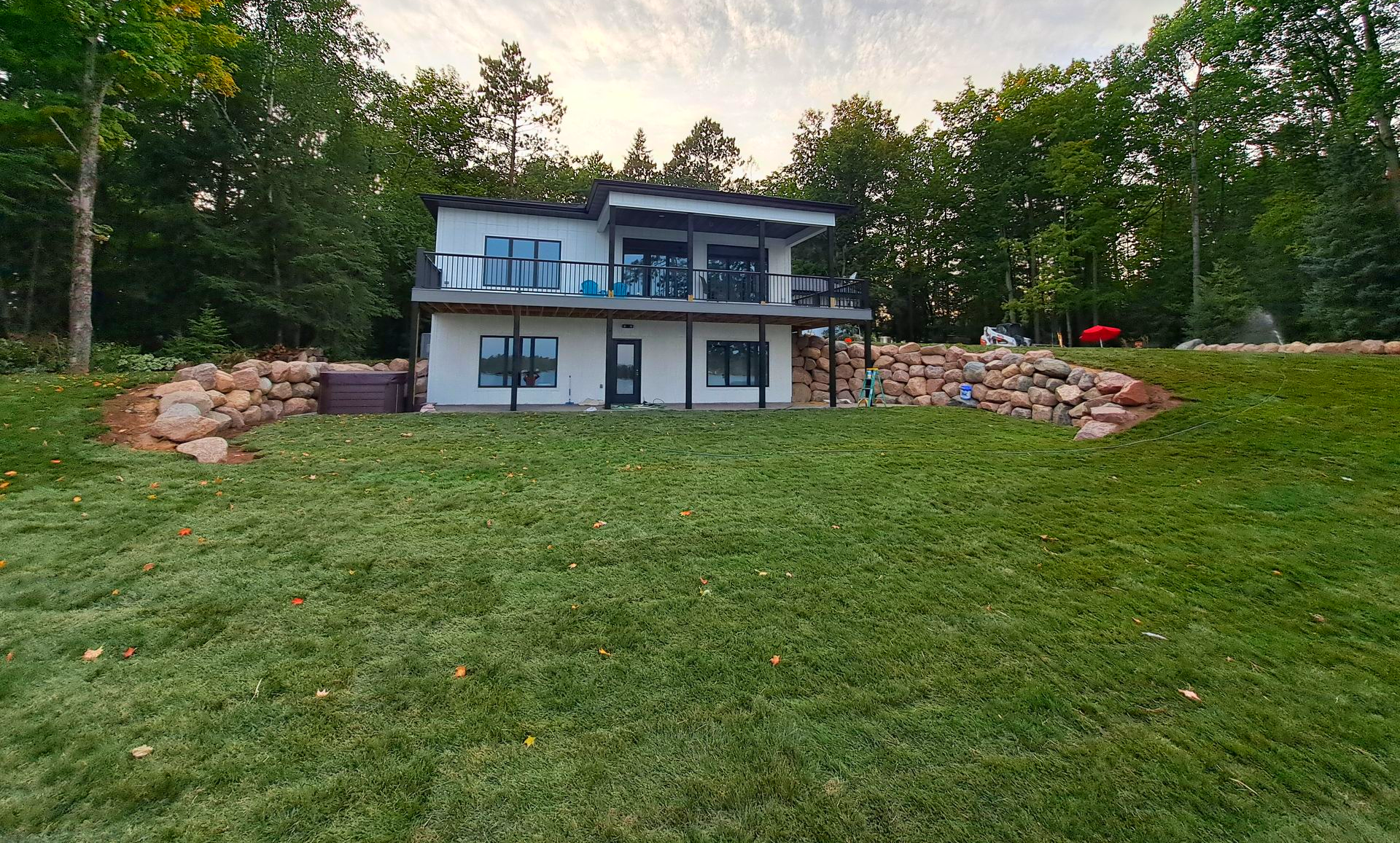 Two-story white house with black trim, balcony, and rock retaining wall on a grassy hill, surrounded by trees.