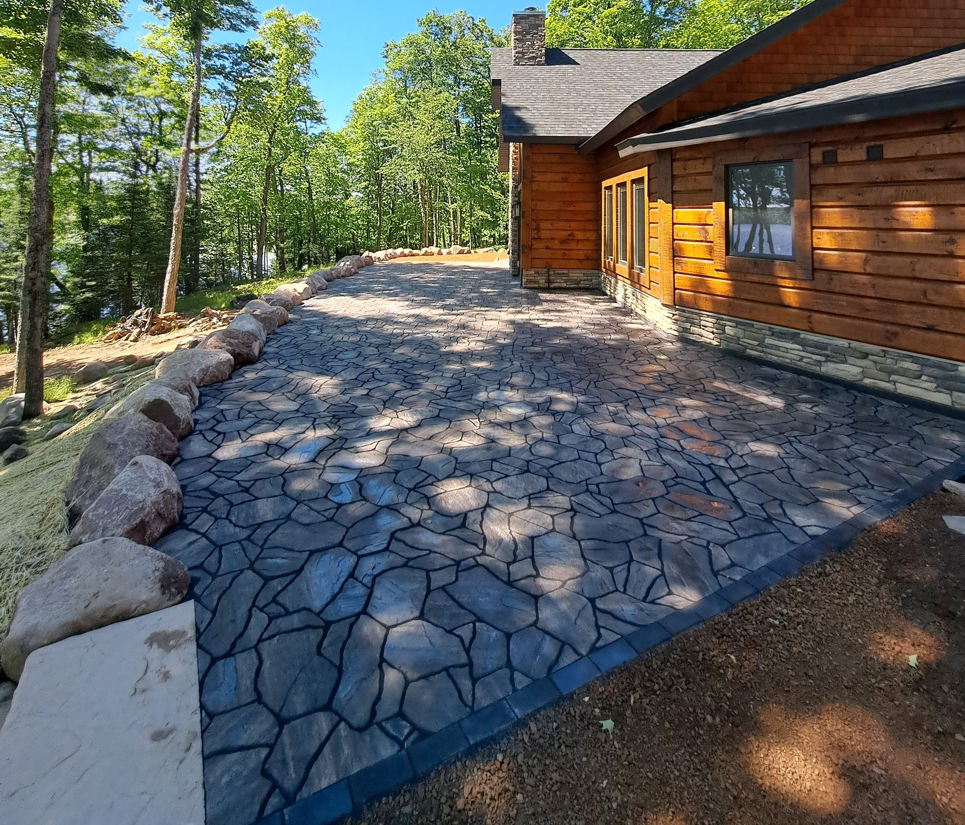 Stone patio next to a log cabin, bordered by rocks and trees; sunny day.