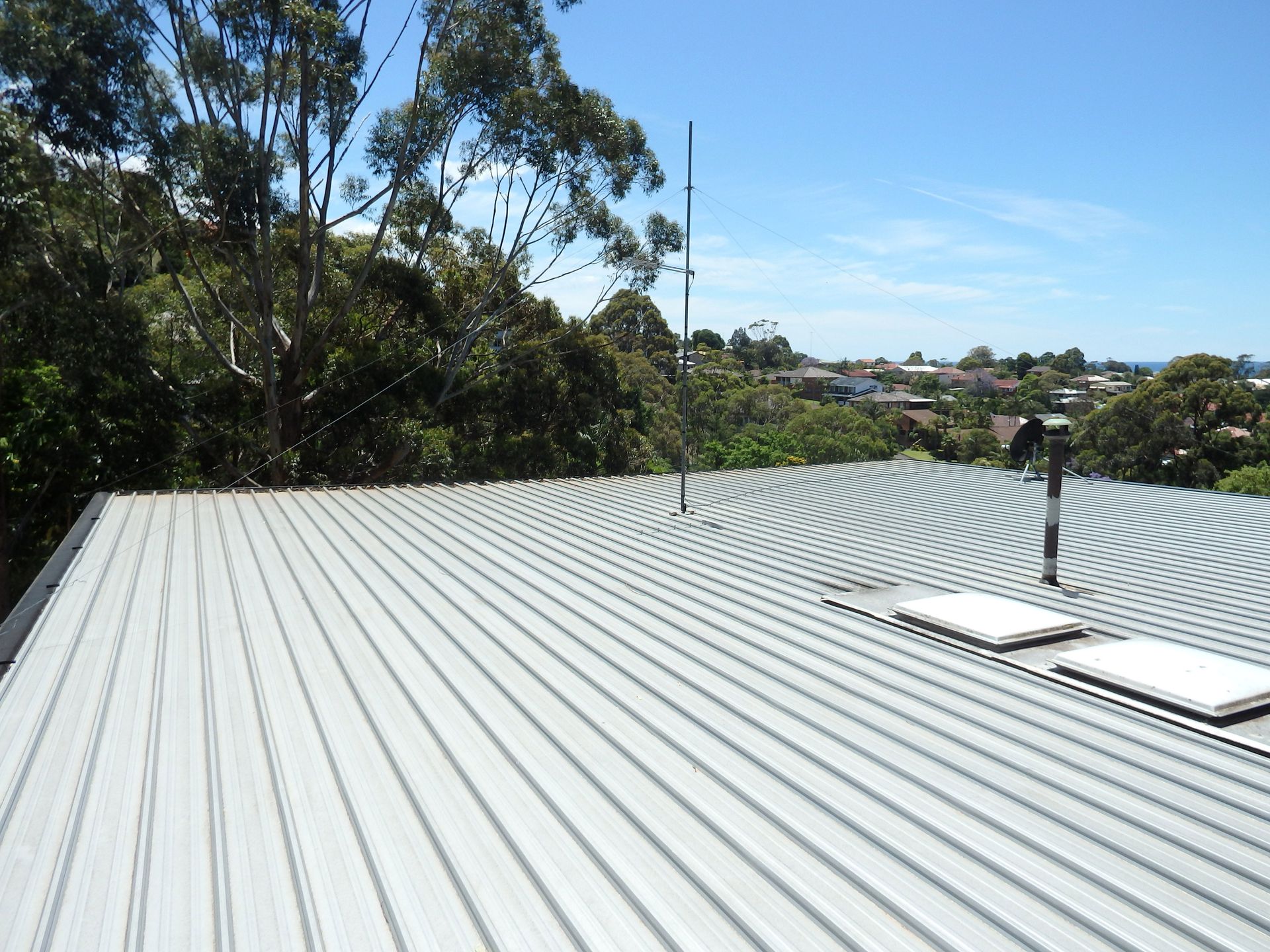 A corrugated metal roof with skylights and an antenna, viewed against a background of trees and houses under a blue sky.
