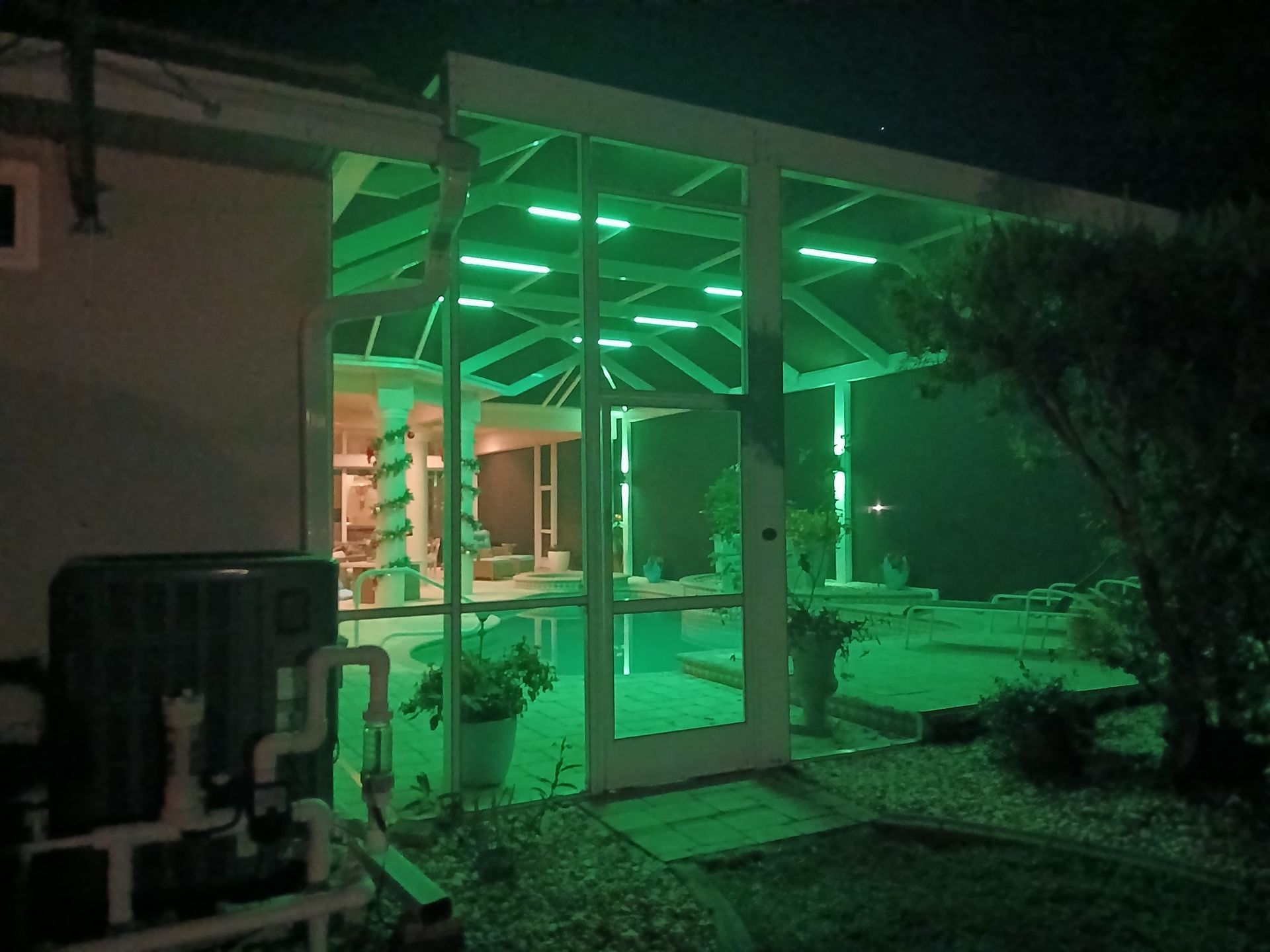 Green-lit pool enclosure at night. A pool, patio, and air conditioning unit are visible.