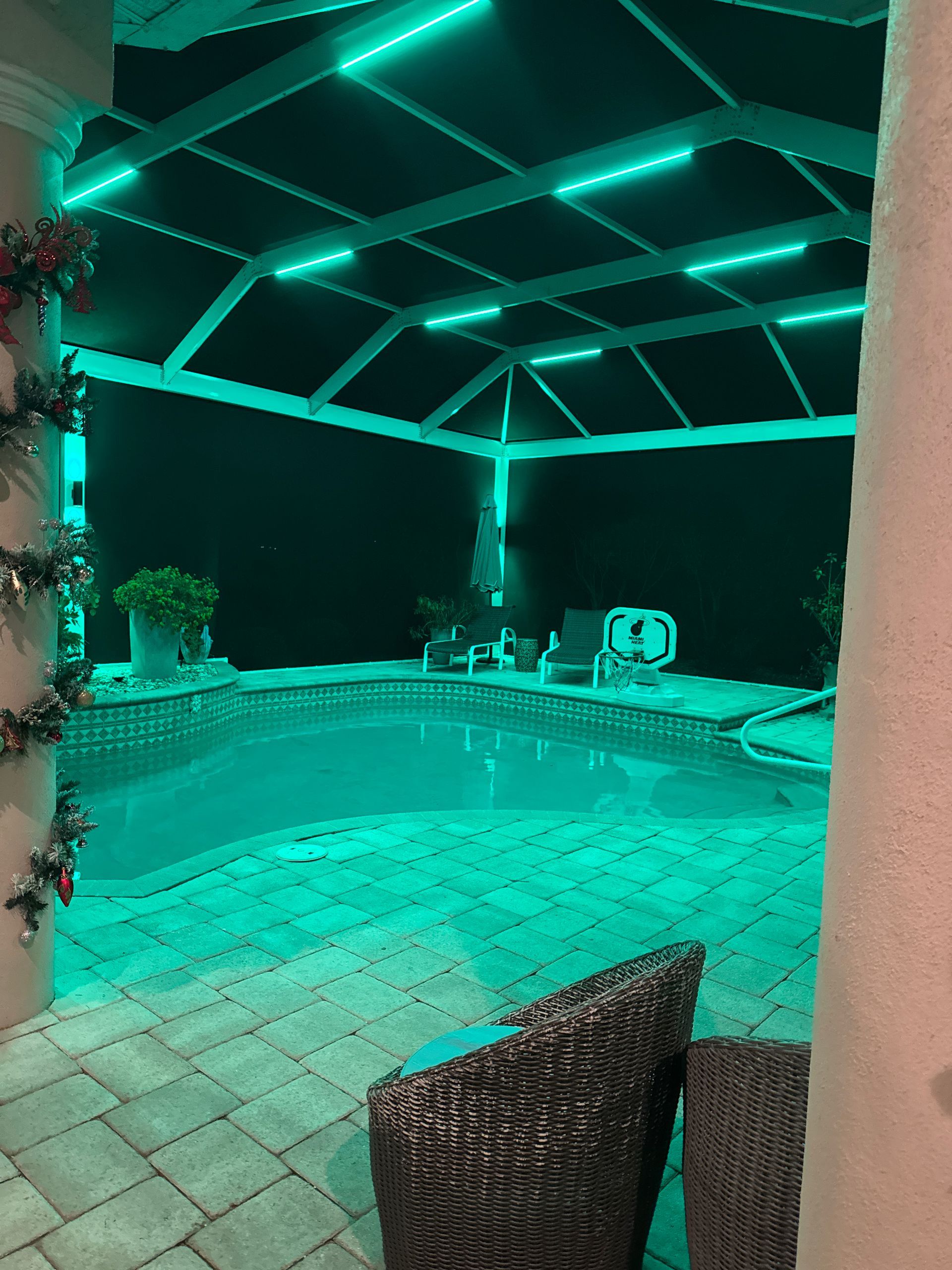 Pool area at night illuminated with green lights. Patio furniture by pool, structure overhead.