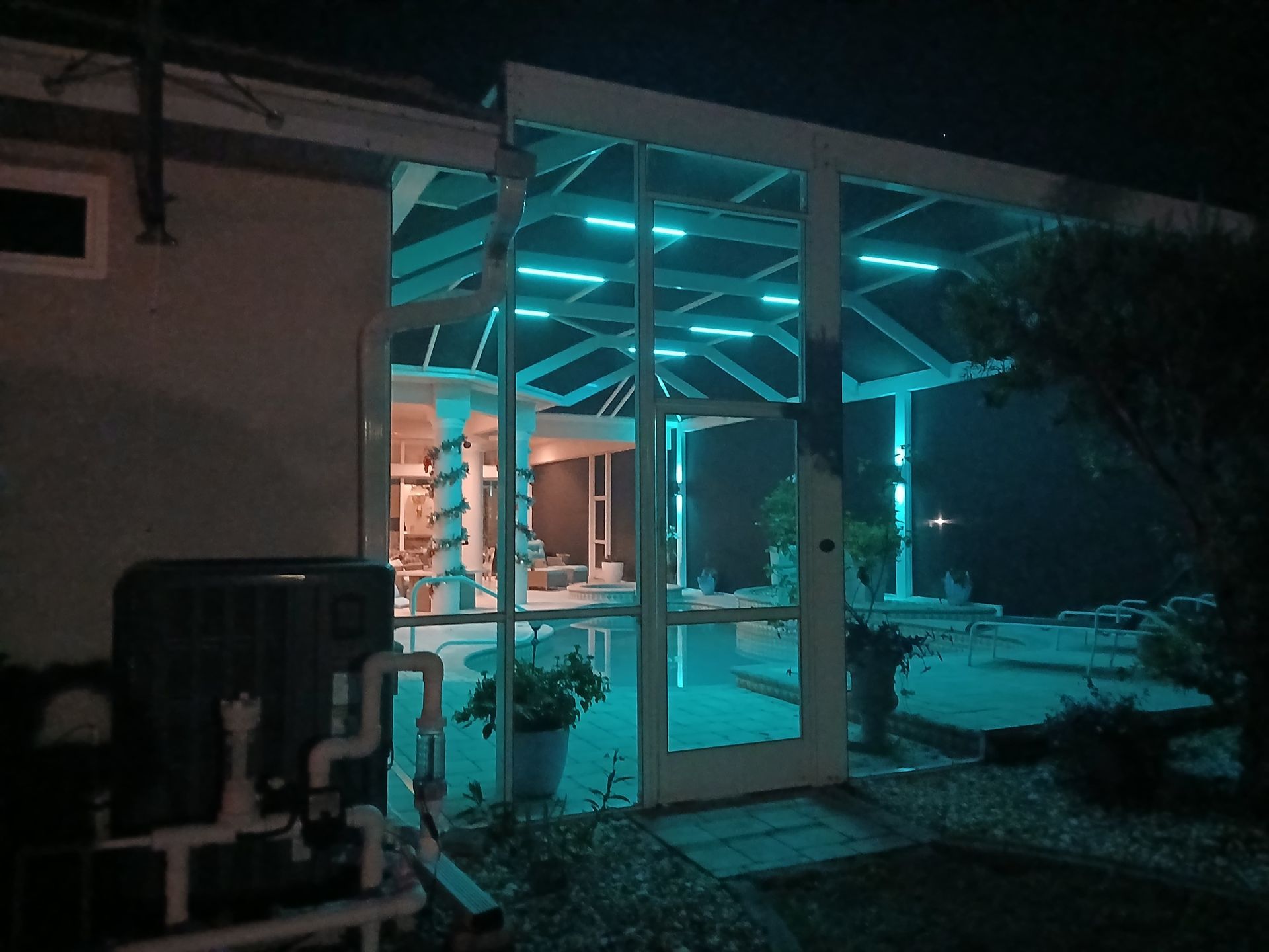 Nighttime view of a backyard pool enclosure illuminated with turquoise lights. Includes pool equipment, plants, and a glimpse of the pool area.
