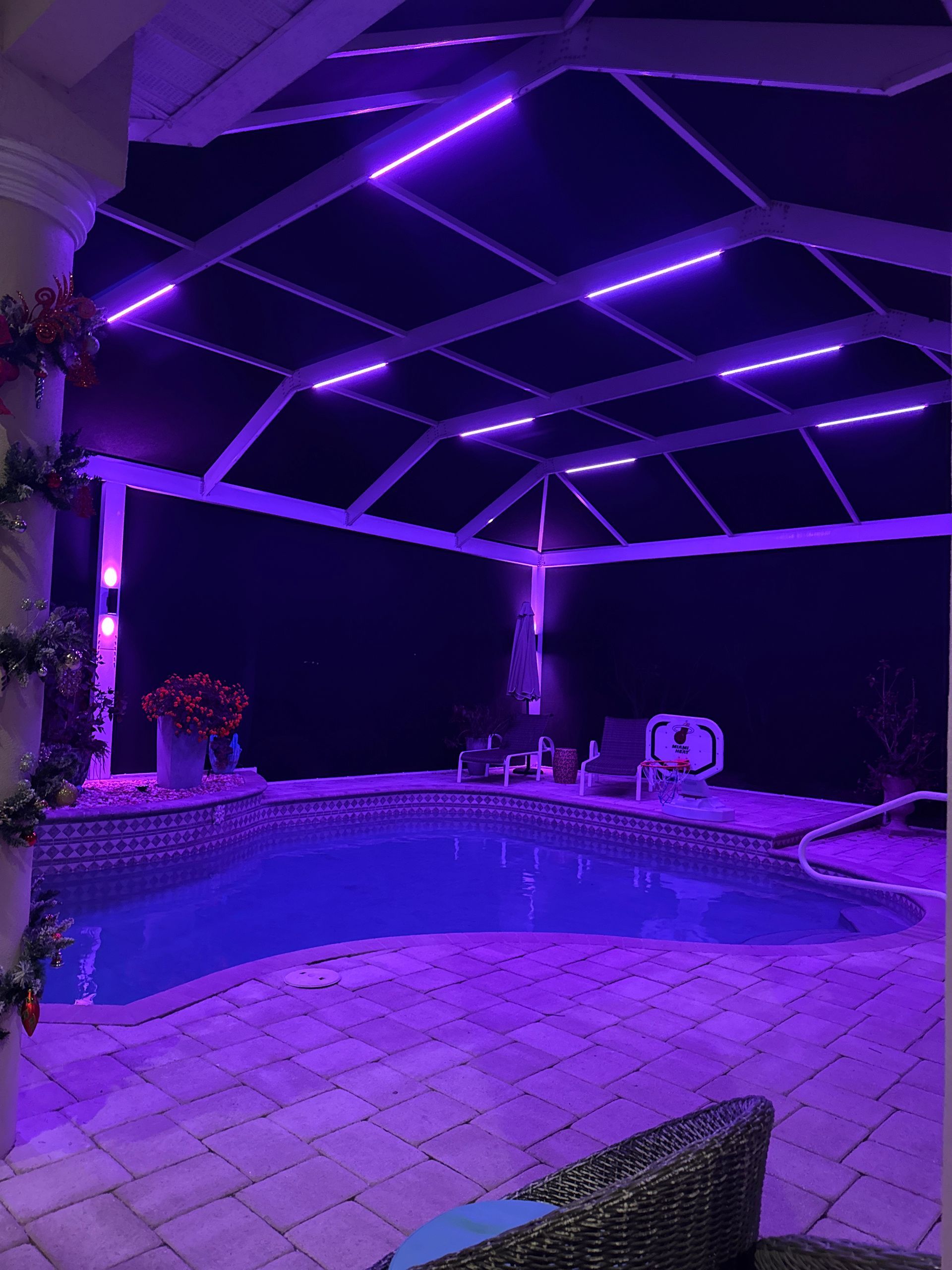 Purple-lit pool area at night. Screened enclosure with overhead lights. Pool is blue, with a chair and plants.