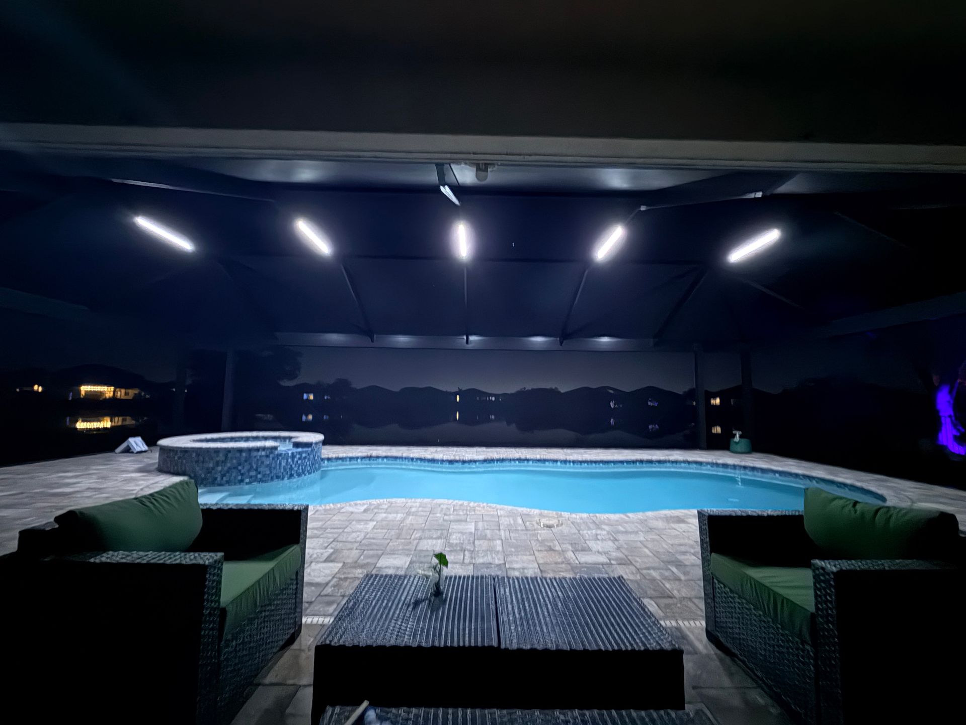 Night view of a pool with seating area under a canopy. Pool and hot tub in blue; green cushions.