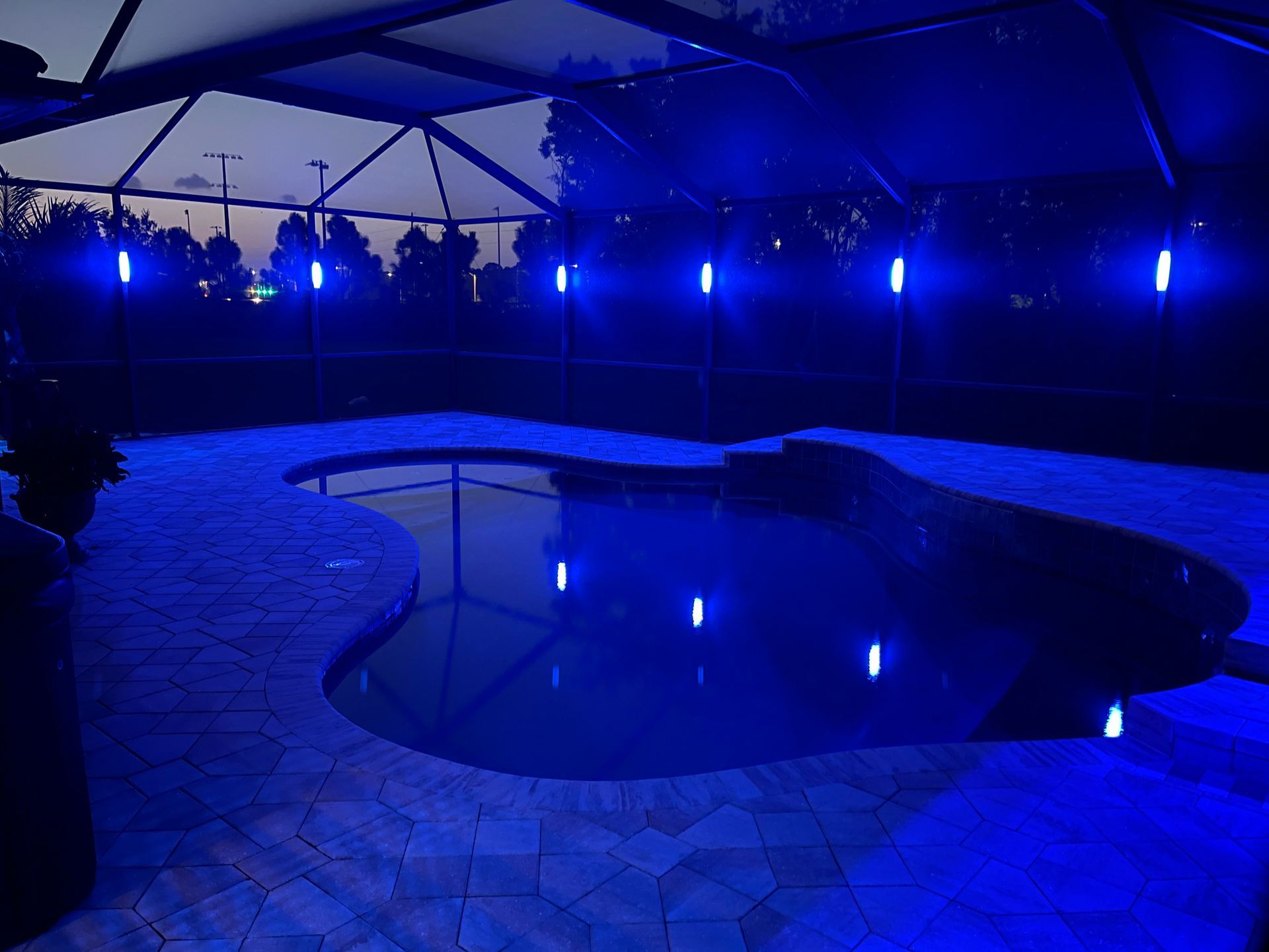 Blue-lit pool at night with tiki torches lining the edge and their reflections in the water. Dark sky in the background.