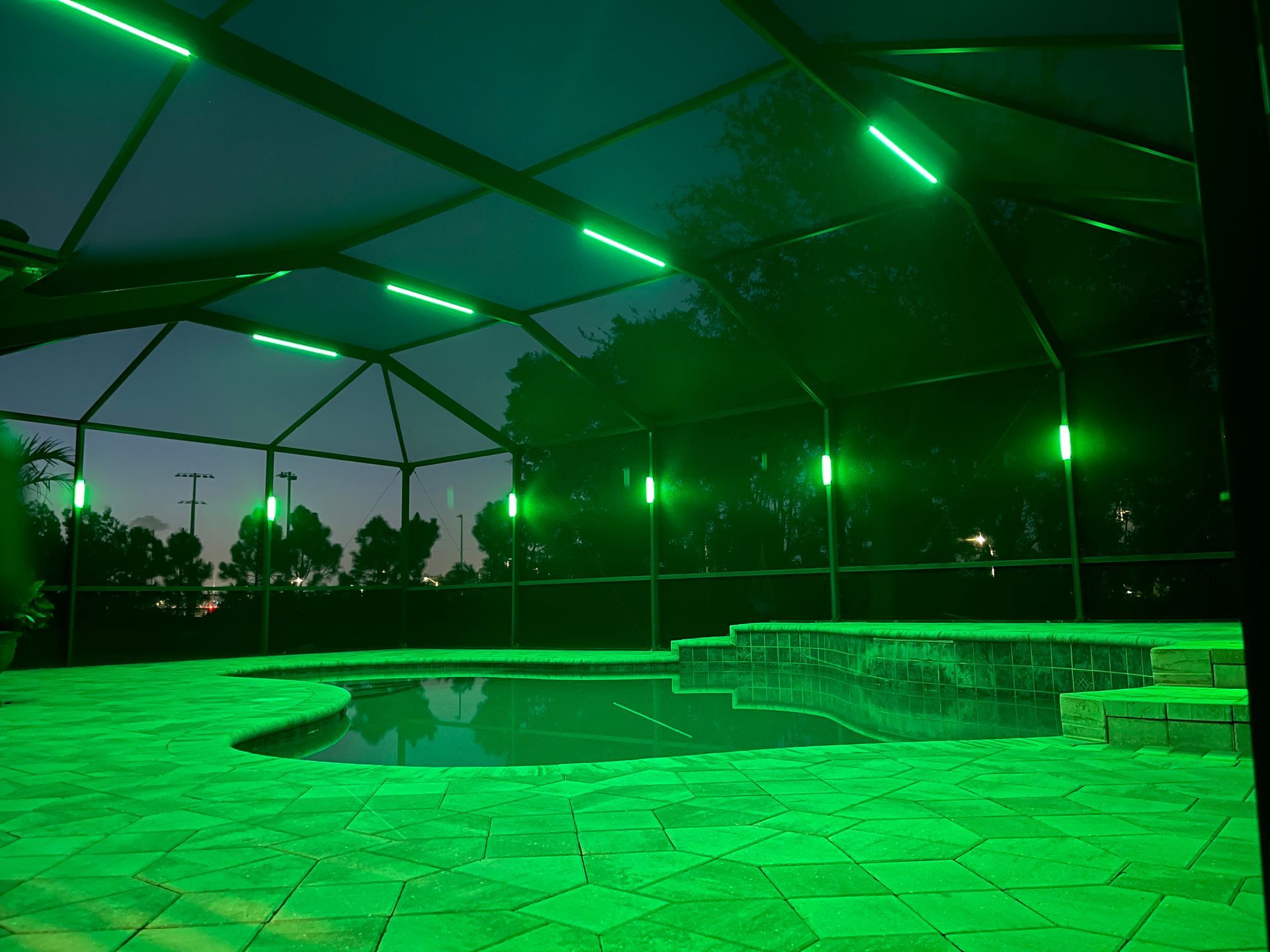 Pool illuminated by green lights at night, under a screened enclosure.