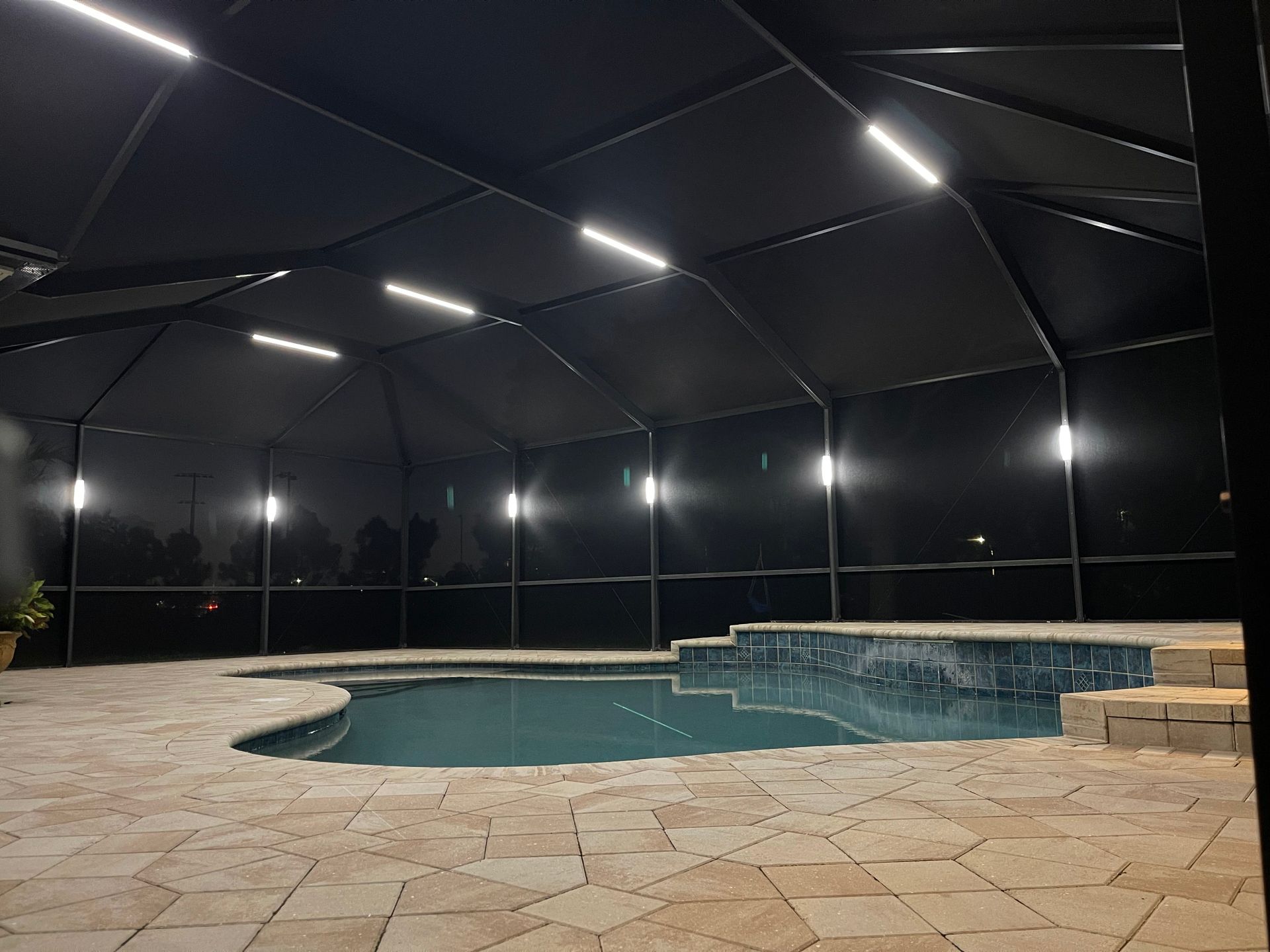 Night view of a swimming pool enclosed in a black screen structure with overhead lighting.