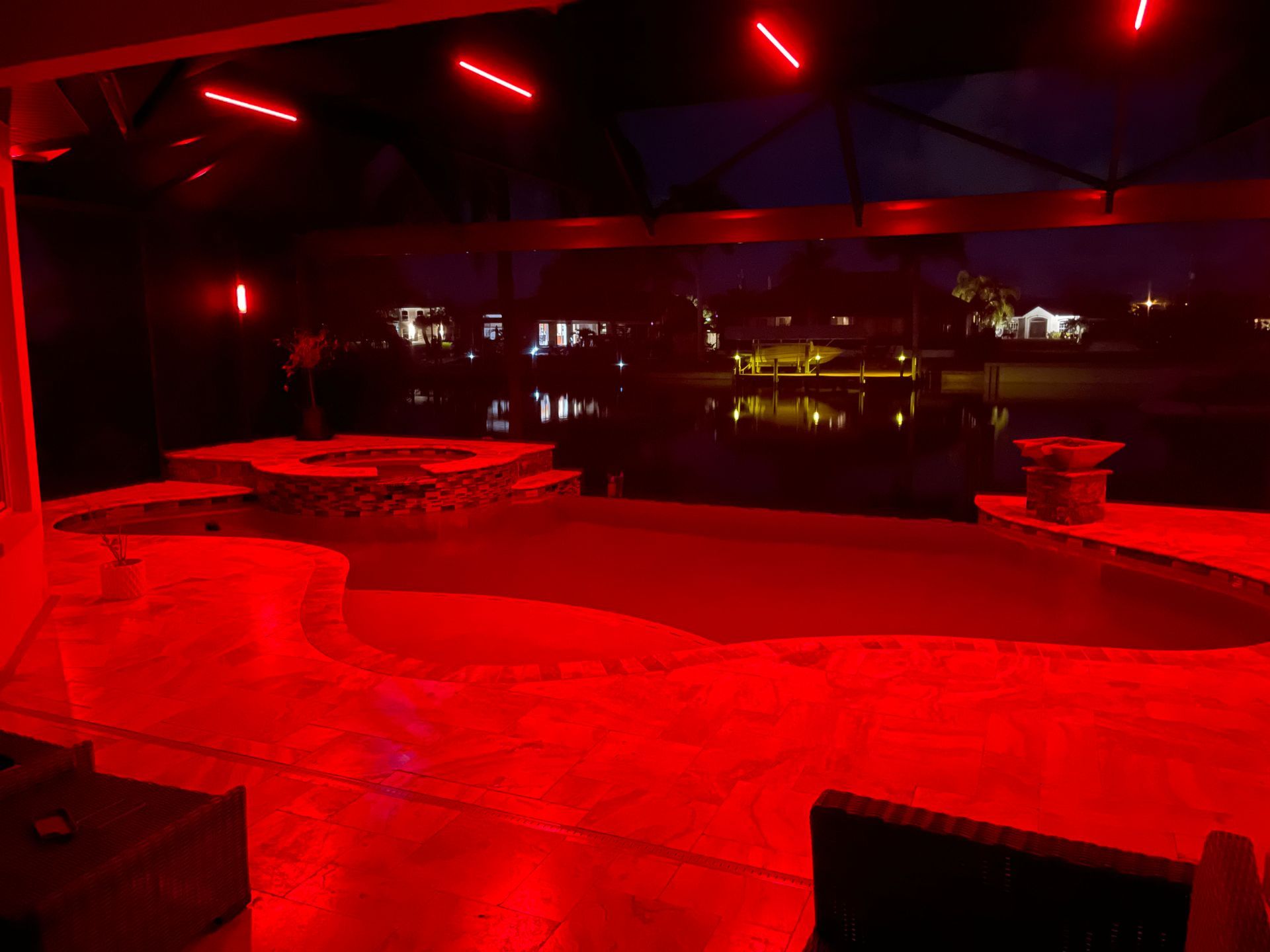 Red-lit pool area at night. Features a pool, spa, and outdoor seating with a view of a body of water and distant lights.