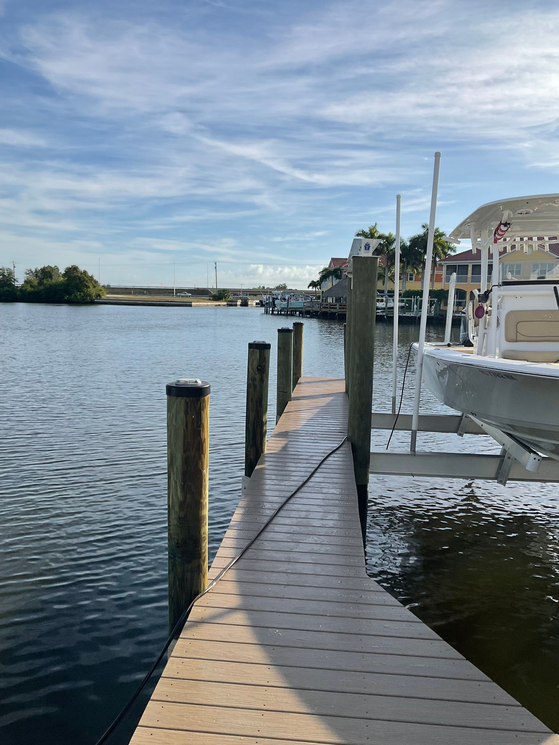 A wooden dock extends into a calm waterway, with boats docked and houses lining the shore, under a blue sky.
