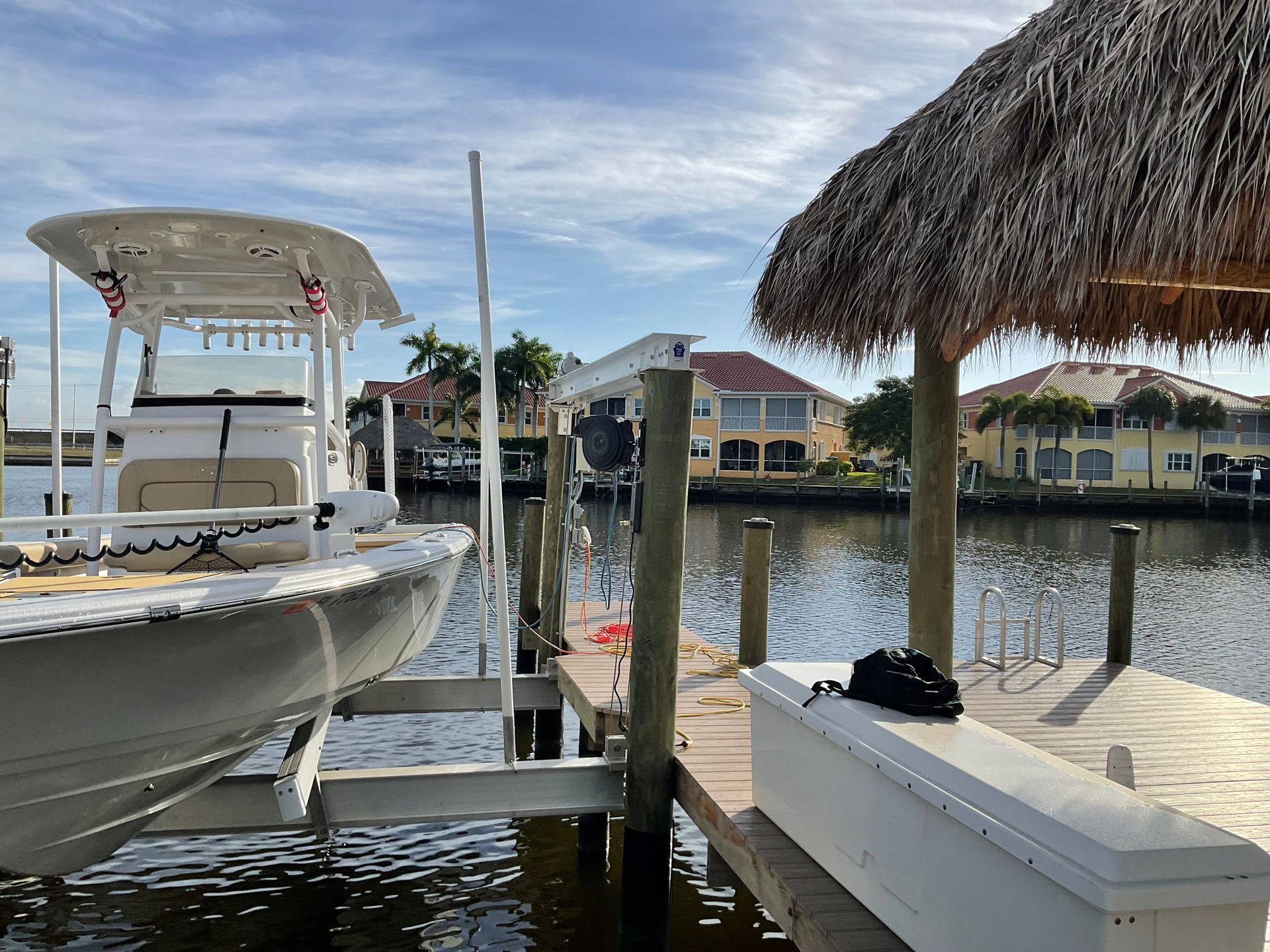 Boat on a lift at a dock with a thatched roof, in front of houses on a canal under a blue sky.
