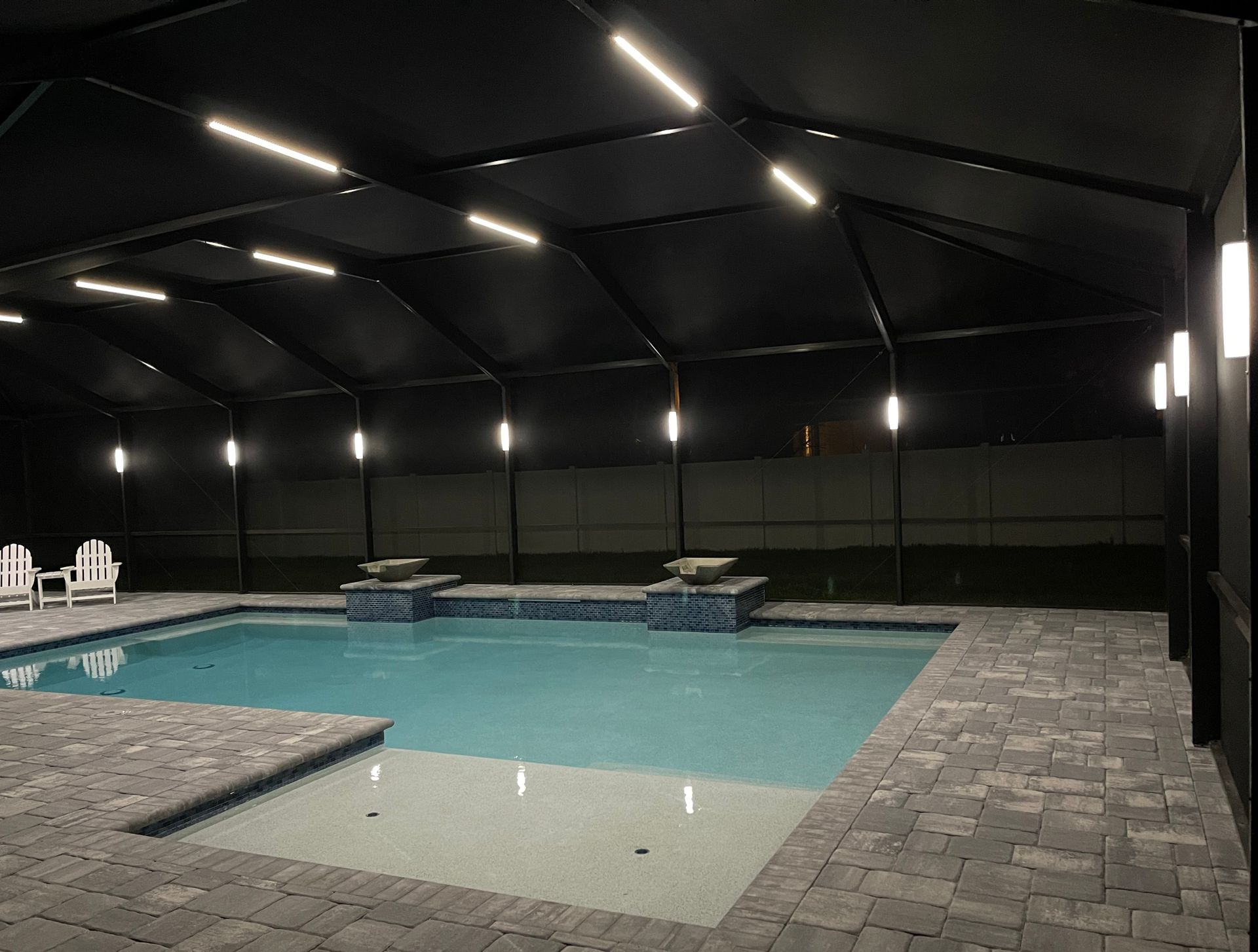 Nighttime view of a screened-in pool and hot tub, lit by overhead and wall lights, against a dark sky.