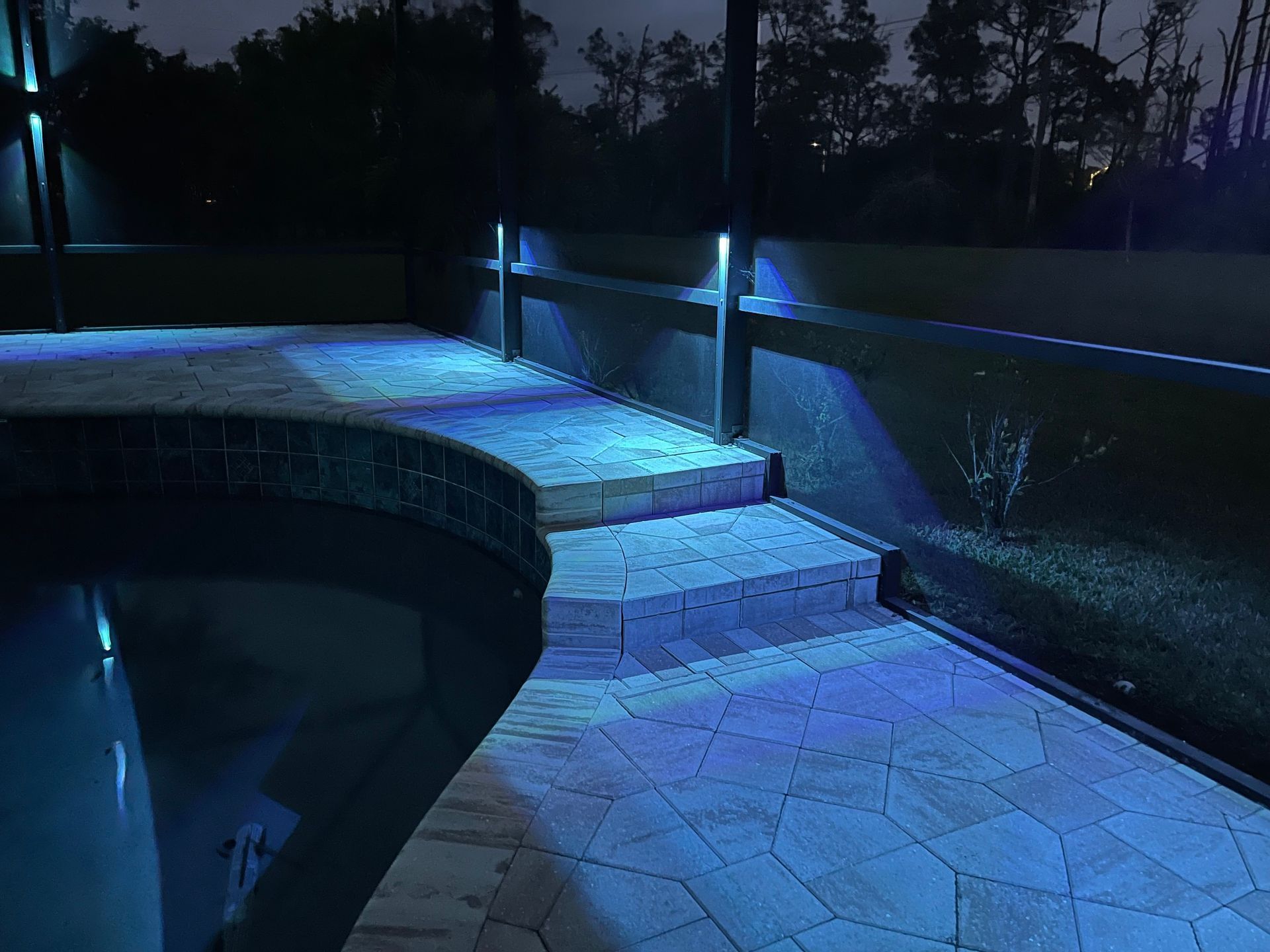 Blue-lit patio at night. Steps lead up to a raised area beside a pool. Small lights illuminate the wall.