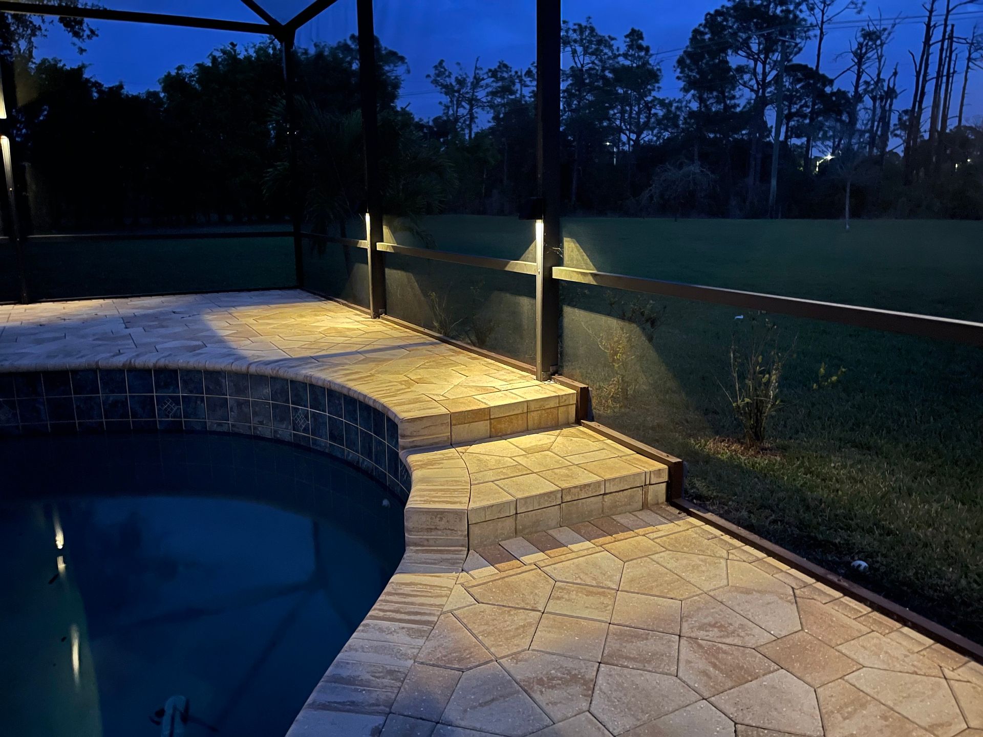 Lit steps and pathway next to a pool at dusk, with ambient lighting on the fence.
